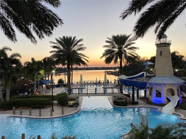 pool at dusk featuring a water slide, a community pool, a water view, and a patio area