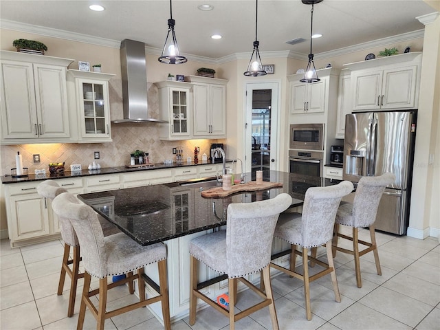 kitchen with stainless steel appliances, wall chimney range hood, an island with sink, dark stone counters, and ornamental molding