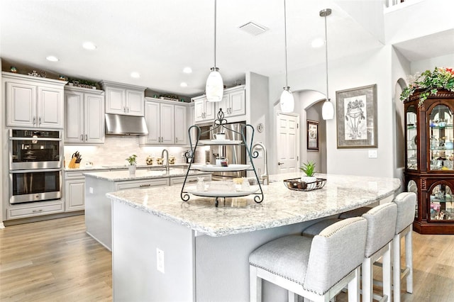 kitchen with light stone counters, hanging light fixtures, a kitchen island with sink, appliances with stainless steel finishes, and light wood-type flooring