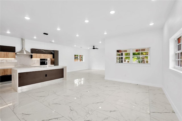 kitchen with decorative light fixtures, wall chimney exhaust hood, tasteful backsplash, an island with sink, and ceiling fan