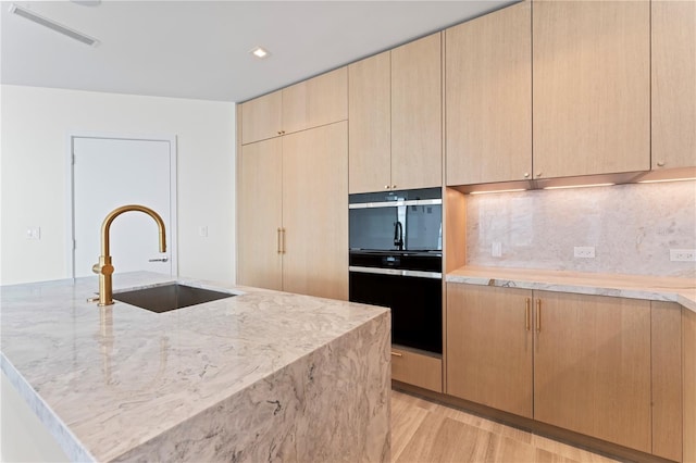 kitchen featuring black oven, a kitchen island with sink, light brown cabinetry, light hardwood / wood-style floors, and sink