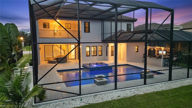 back house at dusk with a swimming pool with hot tub, a balcony, glass enclosure, and a patio area