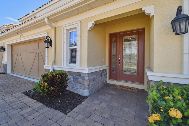 doorway to property featuring an attached garage, stone siding, decorative driveway, and stucco siding