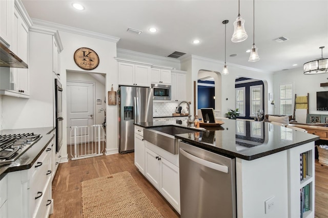 kitchen with appliances with stainless steel finishes, hanging light fixtures, white cabinets, wood-type flooring, and a kitchen island with sink