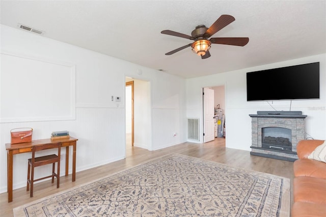 living room with a textured ceiling, ceiling fan, a stone fireplace, and light wood-type flooring