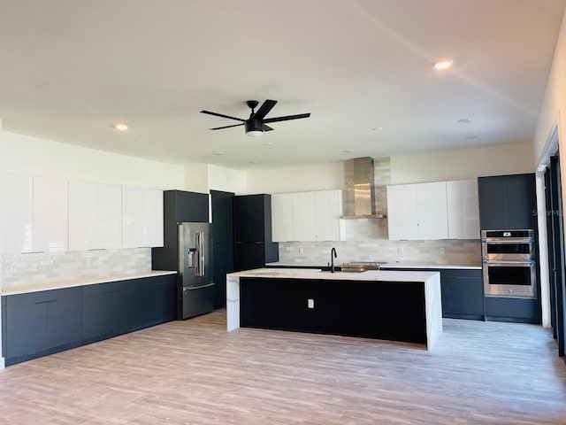 kitchen with a center island with sink, light hardwood / wood-style flooring, wall chimney exhaust hood, white cabinetry, and stainless steel appliances