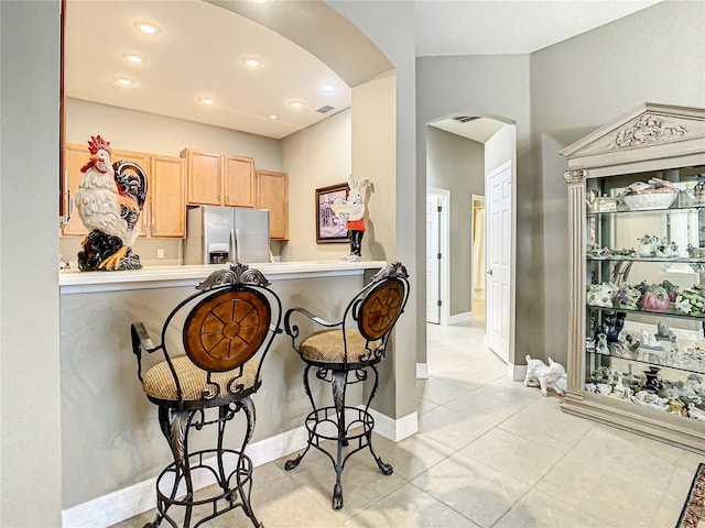 kitchen with stainless steel fridge, light brown cabinets, kitchen peninsula, light tile patterned flooring, and a kitchen bar