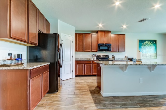 kitchen with a breakfast bar area, a center island with sink, black appliances, light stone counters, and light hardwood / wood-style floors