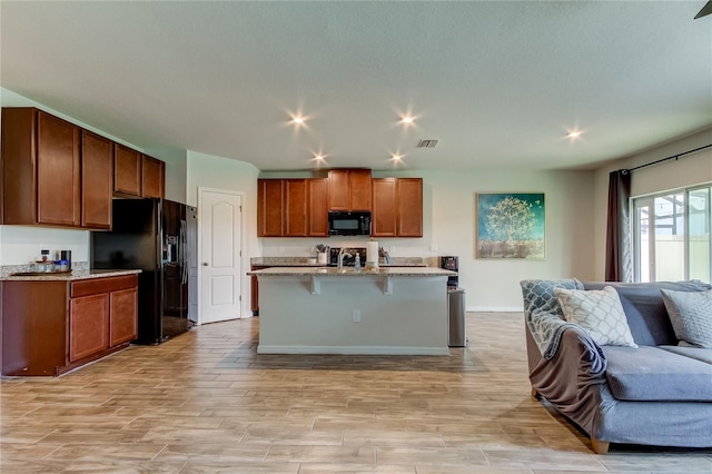 kitchen featuring a breakfast bar area, an island with sink, black appliances, light stone counters, and light hardwood / wood-style floors