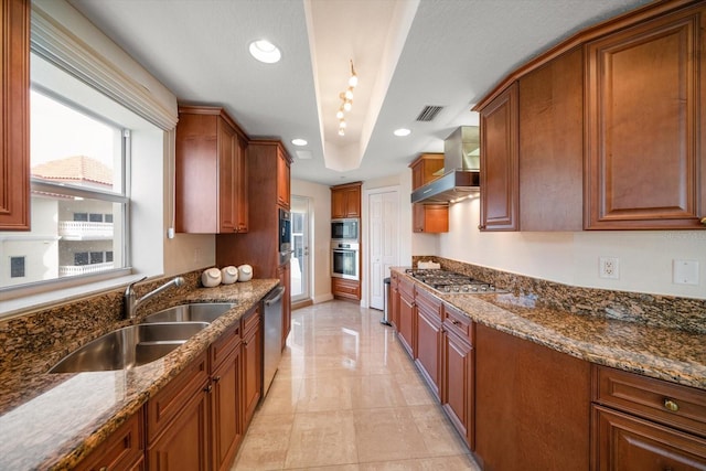 kitchen with brown cabinets, appliances with stainless steel finishes, a sink, wall chimney range hood, and dark stone counters