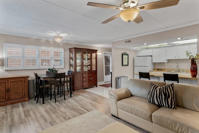 living room featuring a ceiling fan, visible vents, light wood finished floors, ornamental molding, and a textured ceiling