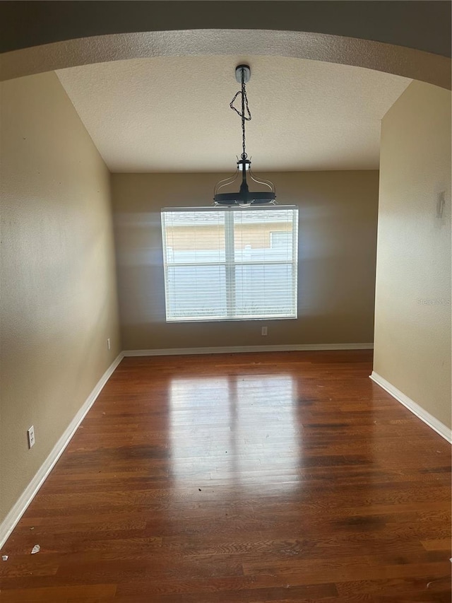 unfurnished dining area with arched walkways, a textured ceiling, baseboards, and wood finished floors