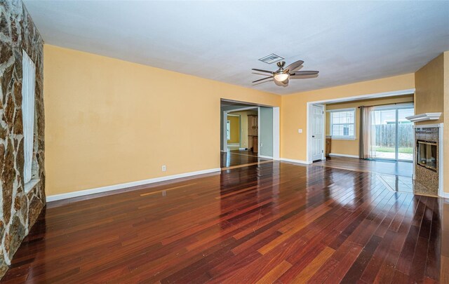 unfurnished living room with ceiling fan, a stone fireplace, and dark wood-type flooring