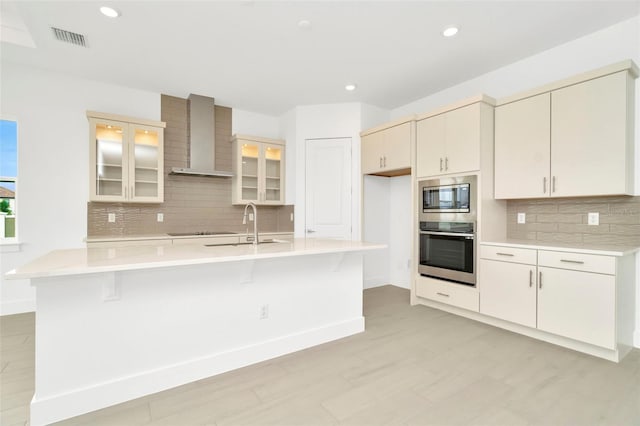 kitchen with visible vents, wall chimney range hood, an island with sink, light countertops, and stainless steel appliances