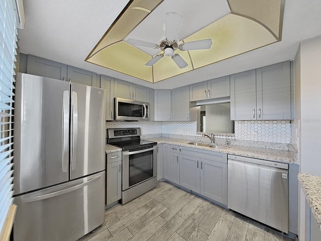 kitchen with light wood-type flooring, appliances with stainless steel finishes, light stone counters, sink, and ceiling fan