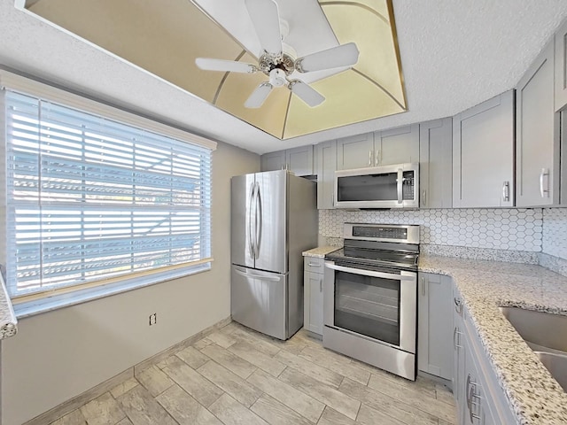 kitchen featuring gray cabinets, backsplash, light stone countertops, ceiling fan, and appliances with stainless steel finishes