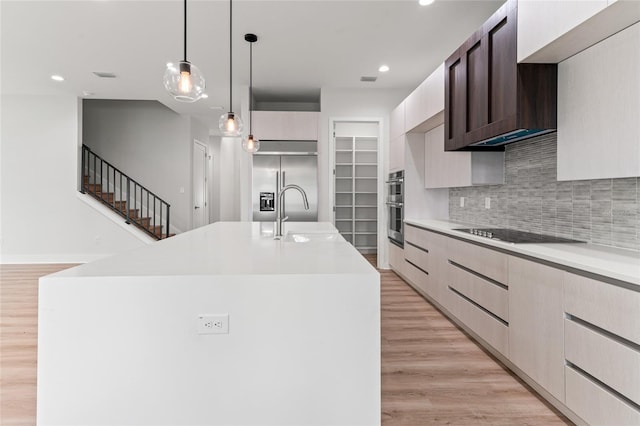 kitchen with a kitchen island with sink, appliances with stainless steel finishes, hanging light fixtures, light hardwood / wood-style floors, and decorative backsplash