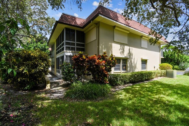 view of side of home featuring stucco siding, a shingled roof, a lawn, a sunroom, and mansard roof