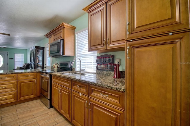 kitchen with a textured ceiling, stainless steel appliances, sink, light wood-type flooring, and dark stone counters