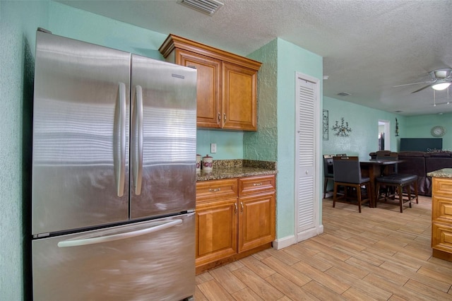 kitchen with dark stone countertops, light wood-type flooring, stainless steel refrigerator, ceiling fan, and a textured ceiling