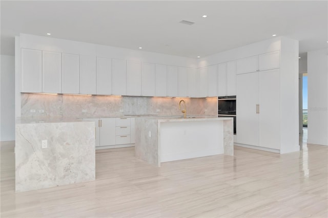 kitchen with white cabinets, light hardwood / wood-style flooring, sink, a center island with sink, and tasteful backsplash