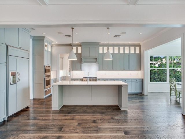 kitchen featuring an island with sink, paneled built in fridge, hanging light fixtures, decorative backsplash, and dark hardwood / wood-style flooring