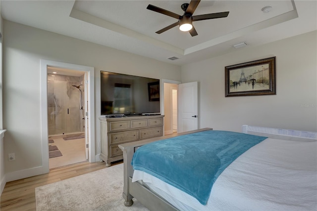 bedroom with light wood-type flooring, a tray ceiling, ceiling fan, and ensuite bath