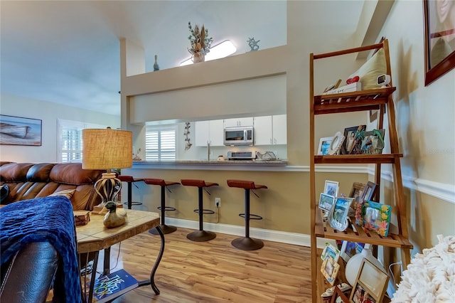 interior space featuring stainless steel microwave, a kitchen breakfast bar, wood finished floors, white cabinetry, and a towering ceiling