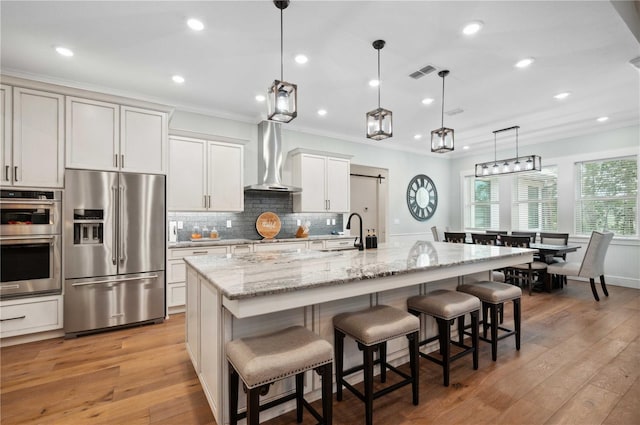 kitchen featuring wall chimney exhaust hood, hanging light fixtures, stainless steel appliances, light hardwood / wood-style flooring, and a spacious island