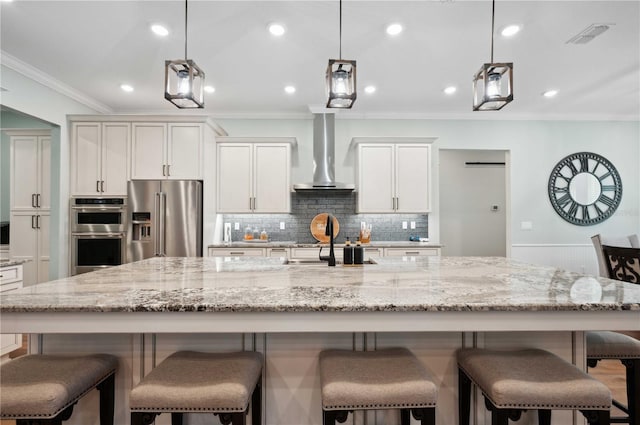 kitchen featuring a large island with sink, white cabinets, wall chimney range hood, appliances with stainless steel finishes, and decorative light fixtures