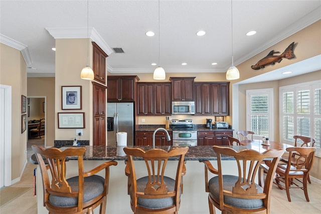 kitchen featuring light tile patterned floors, appliances with stainless steel finishes, a kitchen island with sink, hanging light fixtures, and dark stone counters