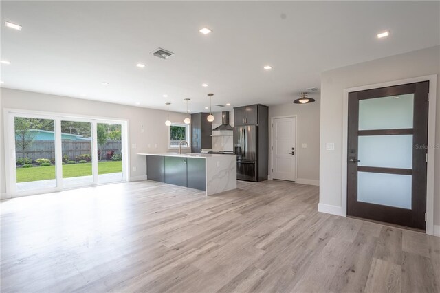 kitchen with hanging light fixtures, stainless steel fridge, a center island with sink, wall chimney exhaust hood, and light hardwood / wood-style flooring