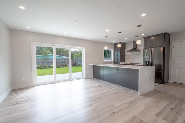 kitchen featuring hanging light fixtures, a kitchen island with sink, wall chimney range hood, stainless steel refrigerator with ice dispenser, and light hardwood / wood-style floors