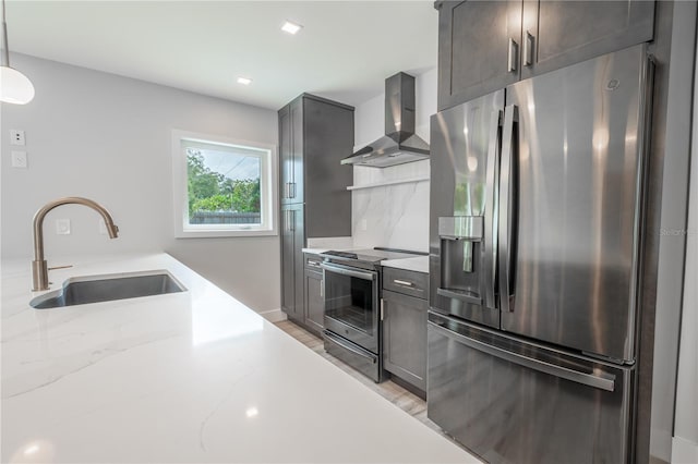 kitchen with light stone countertops, stainless steel appliances, decorative light fixtures, sink, and wall chimney range hood