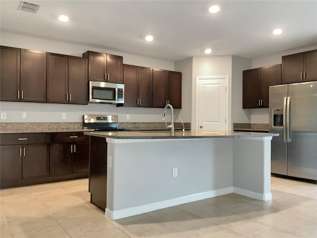 kitchen featuring stainless steel appliances, dark stone countertops, dark brown cabinetry, and a kitchen island with sink