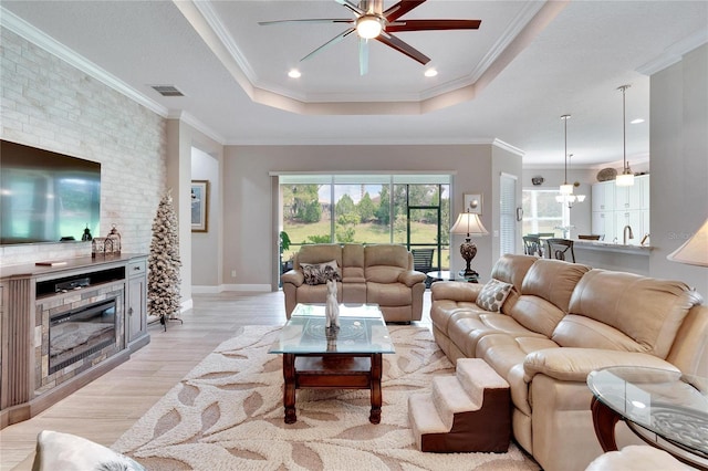 living room featuring a raised ceiling, a fireplace, light wood-style flooring, ceiling fan, and ornamental molding