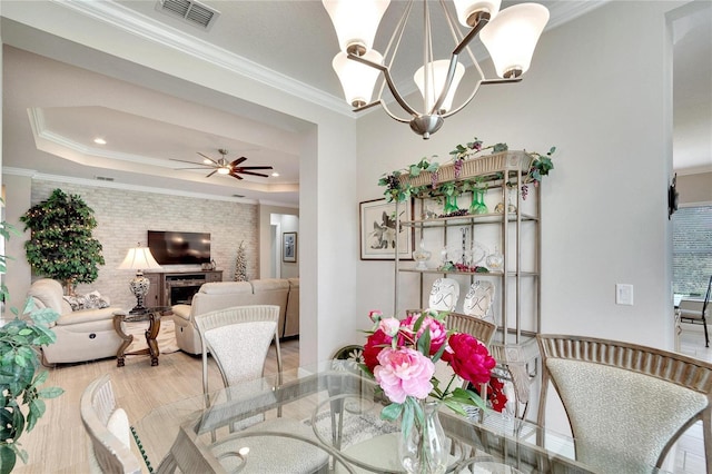 dining area with crown molding, ceiling fan with notable chandelier, a raised ceiling, visible vents, and a brick fireplace