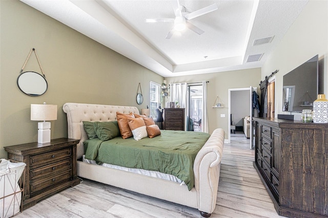 bedroom with a barn door, light hardwood / wood-style floors, a tray ceiling, ceiling fan, and a textured ceiling