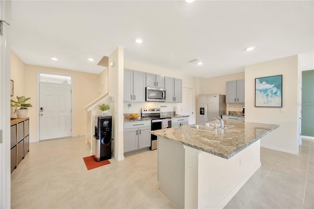 kitchen featuring appliances with stainless steel finishes, light stone countertops, sink, gray cabinets, and a kitchen island with sink