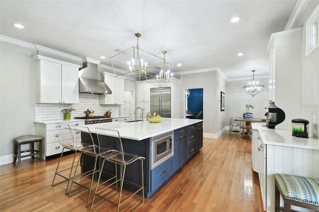 kitchen featuring built in appliances, wall chimney exhaust hood, blue cabinets, a kitchen bar, and white cabinetry