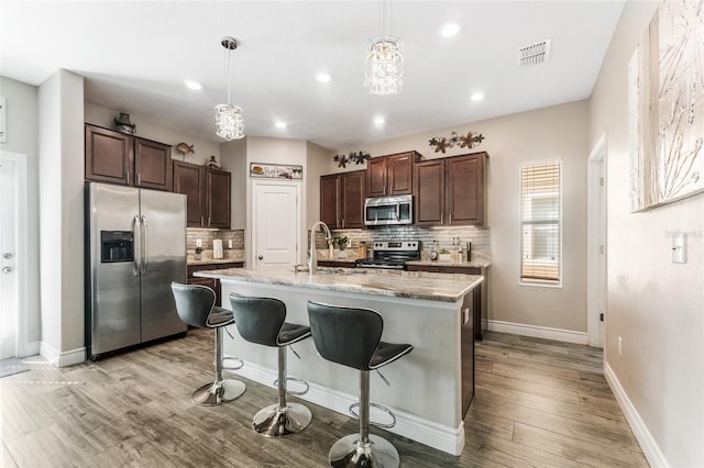 kitchen featuring decorative light fixtures, a kitchen island with sink, backsplash, light hardwood / wood-style flooring, and appliances with stainless steel finishes