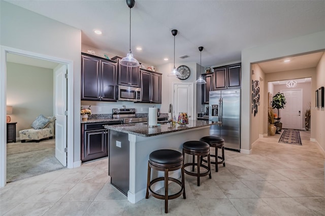 kitchen with a breakfast bar area, appliances with stainless steel finishes, a kitchen island with sink, hanging light fixtures, and dark stone counters