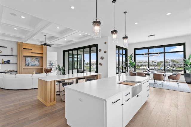 kitchen featuring a kitchen island with sink, coffered ceiling, ceiling fan with notable chandelier, beamed ceiling, and white cabinetry