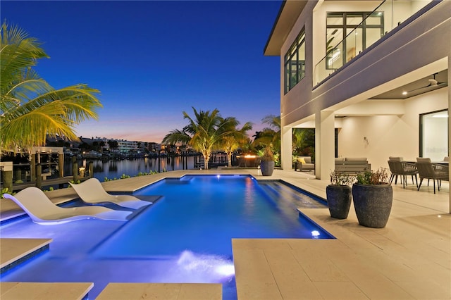 pool at dusk with pool water feature, a water view, and a patio