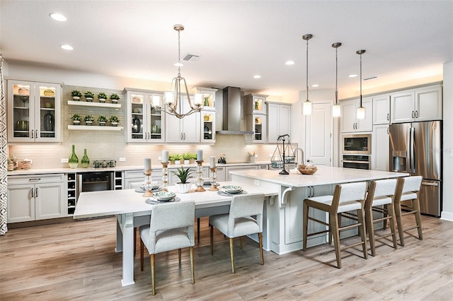 kitchen with wall chimney range hood, light hardwood / wood-style flooring, hanging light fixtures, a large island, and appliances with stainless steel finishes