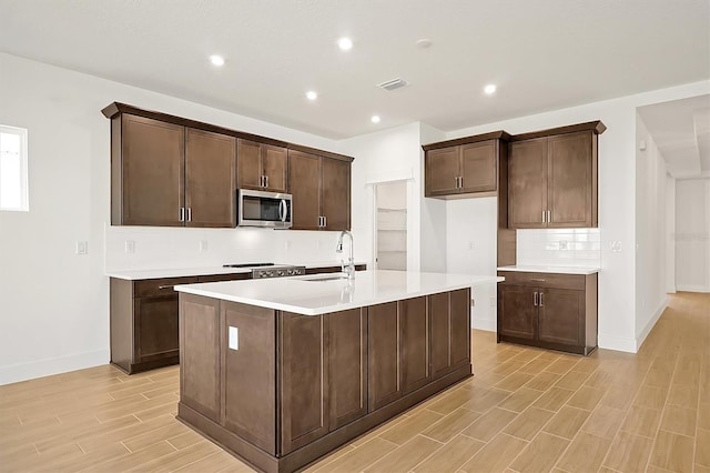 kitchen with stove, backsplash, dark brown cabinetry, sink, and a center island with sink