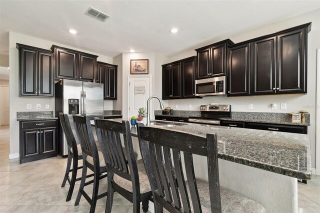 kitchen featuring an island with sink, dark stone countertops, appliances with stainless steel finishes, and a kitchen bar