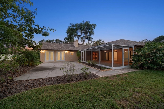 back of house at dusk featuring glass enclosure, french doors, a chimney, a patio area, and a lawn
