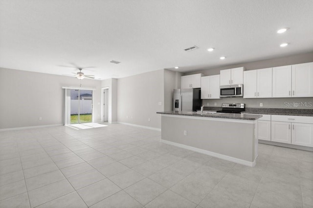 kitchen with appliances with stainless steel finishes, white cabinets, an island with sink, ceiling fan, and dark stone counters