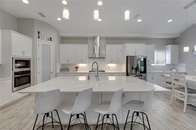 kitchen featuring wall chimney range hood, appliances with stainless steel finishes, pendant lighting, and an island with sink